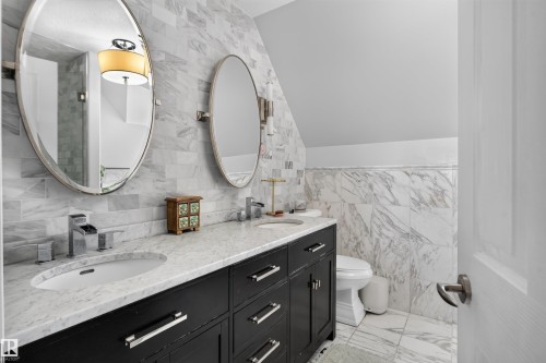 Dual oval mirrors, polished chrome fixtures, black double vanity with drawer storage, stone countertop, and full-height stone tile walls - 4471 Mccrae Avenue Nw, Edmonton, AB - Indoor Photo Showing Bathroom