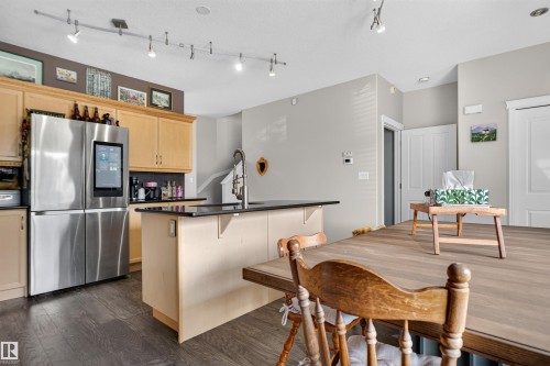Kitchen featuring wood-finish flooring, track lighting, light wood cabinetry, a dark countertop island with a built-in sink, and stainless steel appliances - 4471 Mccrae Avenue Nw, Edmonton, AB - Indoor