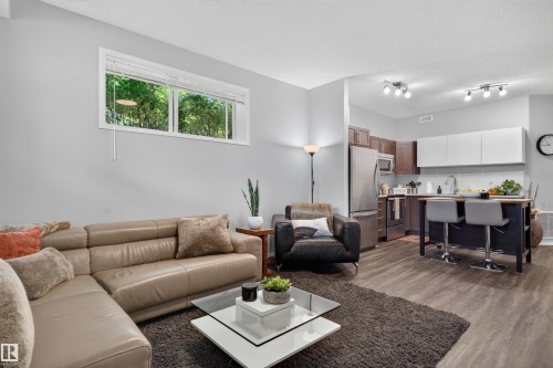 Open-concept living area with wood-finish flooring, a contemporary kitchen featuring stainless steel appliances, dark wood-finish cabinetry, and white overhead cabinetry - #34 6520 2 Av Sw, Edmonton, AB - Indoor Photo Showing Living Room