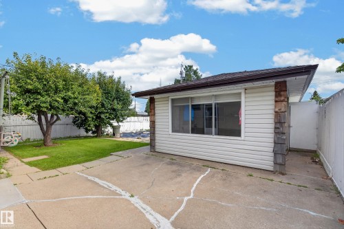 Detached structure with white siding and a brown shingled roof, featuring a large window, concrete patio, and a grassy area with mature trees - 8411 136 Avenue Nw, Edmonton, AB - Outdoor