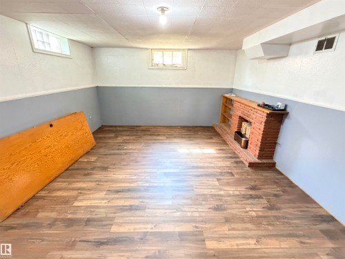 Wood-finish flooring with a brick fireplace, built-in shelving, glass block window, and exposed ceiling light fixture - 10416 100 Avenue, Westlock, AB - Indoor Photo Showing Other Room