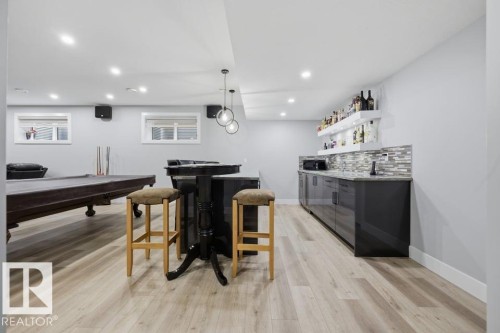 Finished basement recreation space featuring wood-finish flooring, recessed lighting throughout, black cabinetry with mosaic tile backsplash, open shelving, and a wet bar with bar seating - 3303 Soleil Boulevard, Beaumont, AB - Indoor