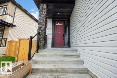 Red entry door with transom window, stone column accent, concrete steps, black railing, and light-colored siding - 3303 Soleil Boulevard, Beaumont, AB - Outdoor With Exterior