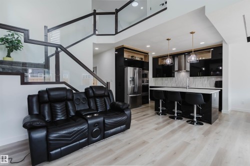 Open-concept living area featuring light wood-finish flooring, a modern glass and dark wood railing, a kitchen island with a white countertop, and sleek black cabinetry with integrated stainless steel appliances - 5320 Kimball Place Sw, Edmonton, AB - Indoor