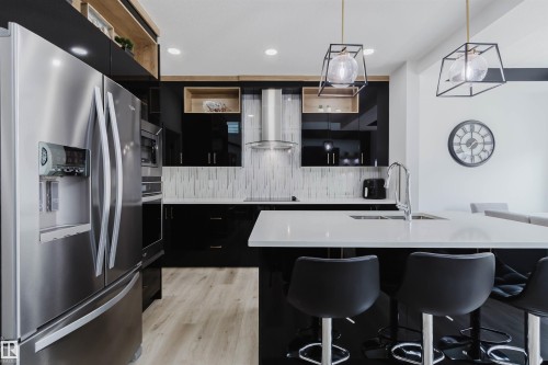 Stainless steel French door refrigerator, black high-gloss cabinetry, wood-finish open shelving, white tile backsplash, and a white countertop island with a built-in sink - 5320 Kimball Place Sw, Edmonton, AB - Indoor Photo Showing Kitchen With Upgraded Kitchen