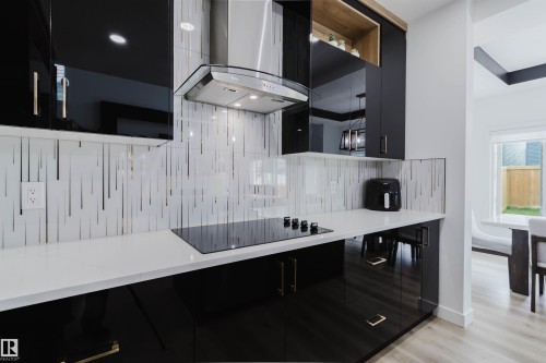 Sleek kitchen featuring black high-gloss cabinetry, integrated cooktop, stainless steel range hood, and white countertops - 5320 Kimball Place Sw, Edmonton, AB - Indoor
