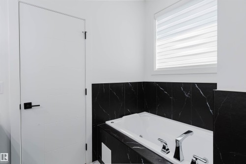 White soaking tub with chrome fixtures, surrounded by black marble-patterned tile, set beneath a window with layered window treatments - 5320 Kimball Place Sw, Edmonton, AB - Indoor Photo Showing Bathroom