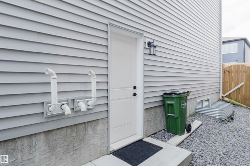 Light gray siding exterior with a white entry door, exterior wall lantern, concrete foundation, and a window well with a metal grate - 5320 Kimball Place Sw, Edmonton, AB - Outdoor With Exterior