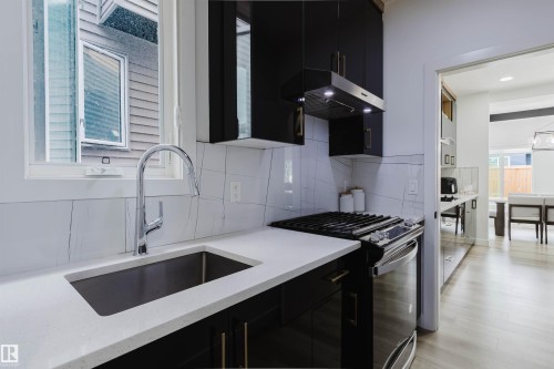 Stainless steel sink with a high-arch faucet, white countertops, black cabinetry with gold-tone hardware, and a gas range with a matching range hood - 5320 Kimball Place Sw, Edmonton, AB - Indoor Photo Showing Kitchen With Upgraded Kitchen