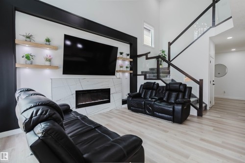 Light wood-finish flooring, a contemporary fireplace with a stone-finish surround, built-in shelving with integrated lighting, and a glass-paneled staircase with dark wood-finish handrails - 5320 Kimball Place Sw, Edmonton, AB - Indoor Photo Showing Living Room With Fireplace