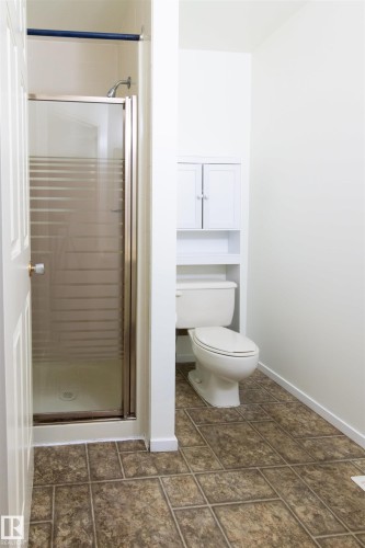 Bathroom featuring a glass-enclosed shower with a chrome showerhead, a white toilet, built-in white cabinetry with a lower open shelf, and textured tile flooring - 4903 11A Avenue Nw, Edmonton, AB - Indoor Photo Showing Bathroom