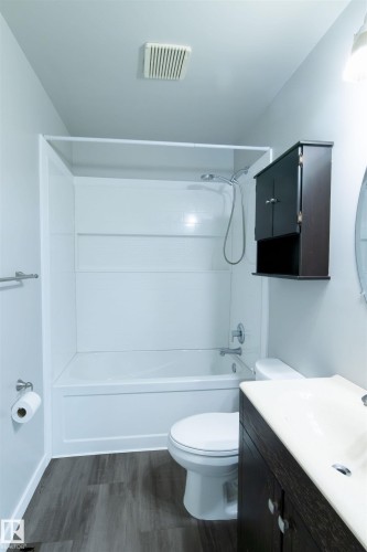Bathroom featuring a white bathtub with shower surround, wood-finish flooring, dark wood vanity with integrated sink, and wall-mounted storage cabinet - 4903 11A Avenue Nw, Edmonton, AB - Indoor Photo Showing Bathroom