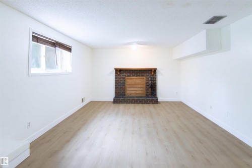 Spacious room featuring a brick fireplace with a wood mantle, light wood-finish flooring, white walls, and a window with blinds - 4903 11A Avenue Nw, Edmonton, AB - Indoor Photo Showing Living Room