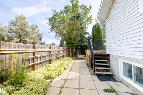 Paved patio area with a wood privacy fence, established perimeter shrubbery, and exterior stairs leading to an elevated deck - 4903 11A Avenue Nw, Edmonton, AB - Outdoor