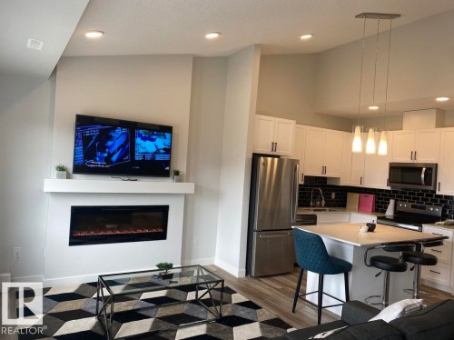 Open-concept living space with vaulted ceilings, a built-in electric fireplace, recessed lighting, and wood-finish flooring - #3 2905 141 St Sw, Edmonton, AB - Indoor Photo Showing Kitchen With Fireplace With Stainless Steel Kitchen