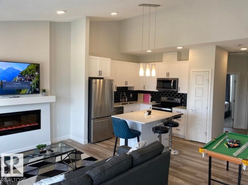 Open-concept living area featuring a modern kitchen with white cabinetry and stainless steel appliances, a built-in fireplace with a white mantel, high vaulted ceilings, and wood-finish flooring - #3 2905 141 St Sw, Edmonton, AB - Indoor Photo Showing Kitchen With Fireplace With Stainless Steel Kitchen