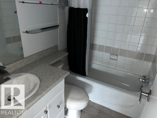 Bathroom featuring a single vanity with an integrated sink, a built-in bathtub with shower, white wall tiling, and wood-finish flooring - #202 9929 113 St Nw, Edmonton, AB - Indoor Photo Showing Bathroom
