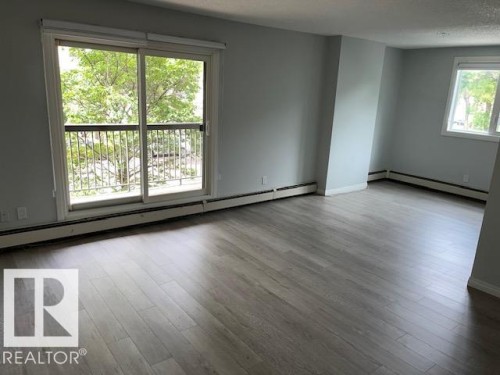Expansive living area with light wood-finish flooring, a large sliding glass door opening to a balcony with metal railing, and a secondary window providing natural light - #202 9929 113 St Nw, Edmonton, AB - Indoor Photo Showing Other Room