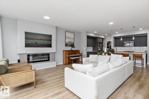 Open-concept living space featuring wood-finish flooring, a built-in electric fireplace, recessed lighting, and a kitchen with dark cabinetry and stainless steel appliances - 3241 Kulay Way Sw, Edmonton, AB - Indoor Photo Showing Living Room With Fireplace