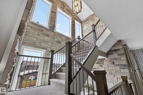 Stone accent wall with multiple windows, carpeted staircase, dark wood railings with metal spindles, and a geometric pendant light fixture - 1399 Graydon Hill Way Sw, Edmonton, AB - Indoor Photo Showing Other Room