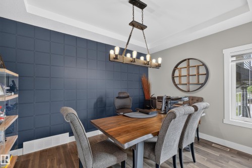 Tray ceiling, wood-finish flooring, accent wall with geometric paneling, natural light from a double-pane window, and a chandelier with Edison-style bulbs - 1399 Graydon Hill Way Sw, Edmonton, AB - Indoor Photo Showing Dining Room