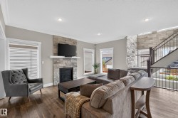 Living room featuring a stone-clad fireplace with a white mantel, wood-finish flooring, recessed lighting, and a multi-level staircase with iron balusters - 