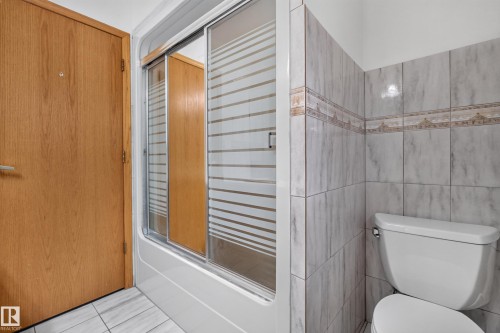 Bathroom featuring a shower-tub combination with sliding glass doors, white toilet, and tiled walls with decorative accent band - 306 Twin Brooks Drive Nw, Edmonton, AB 