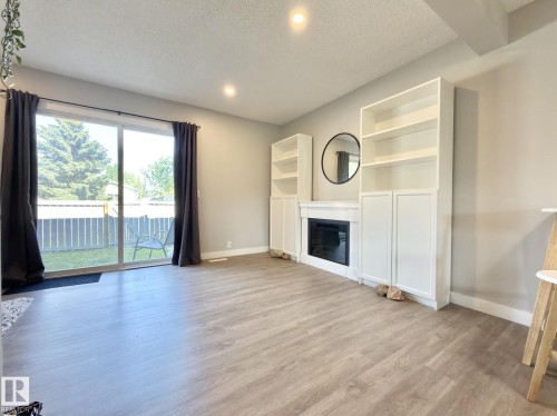 Built-in shelving flanking a central fireplace, wood-finish flooring, recessed lighting, and a sliding glass door leading to a fenced yard - #13 18010 98 Av Nw Nw, Edmonton, AB - Indoor Photo Showing Living Room With Fireplace
