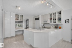 White kitchen with a peninsula, built-in desk, glass-front upper cabinetry, stainless steel sink, and tile flooring - 