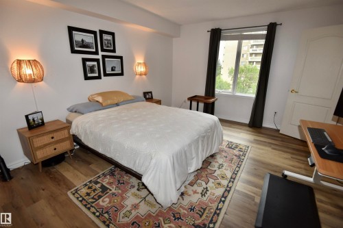 Spacious primary bedroom with hardwood flooring, white bedding, wooden nightstand with integrated lighting, framed wall art, large window with black drapery hardware, and an ornamental area rug anchoring the sleeping area - #706 9741 110 St Nw, Edmonton, AB - Indoor Photo Showing Bedroom