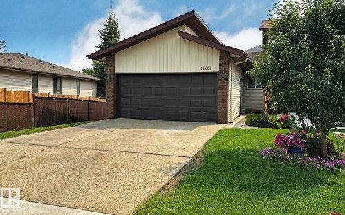 Front exterior with a double-width concrete driveway, integrated garage featuring a dark brown door, and a landscaped lawn area with mature plantings - 11111 157 Avenue Nw, Edmonton, AB - Outdoor With Exterior