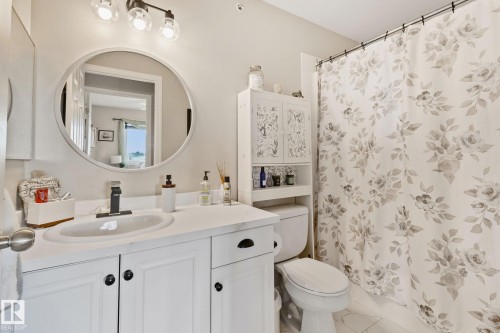 Bathroom featuring a white vanity with a dark faucet, round mirror, and a built-in toilet storage cabinet - #407 4309 33 St, Stony Plain, AB - Indoor Photo Showing Bathroom