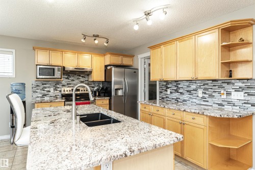 Kitchen featuring a center island with an undermount sink, light wood-finish cabinetry, stainless steel appliances, granite countertops, and a mosaic tile backsplash - 192 Keyport Circle, Leduc, AB - Indoor Photo Showing Kitchen With Double Sink With Upgraded Kitchen