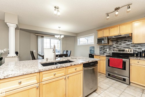Kitchen featuring a granite-topped island with an undermount sink, stainless steel appliances, wood-finish cabinetry, and a mosaic tile backsplash - 192 Keyport Circle, Leduc, AB - Indoor Photo Showing Kitchen With Double Sink