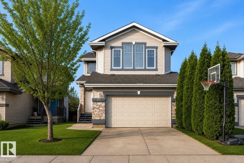 Two-story residential exterior featuring beige siding with dark trim, stone facade accents, an attached garage, a mature front yard tree, and a concrete driveway - 192 Keyport Circle, Leduc, AB - Outdoor With Facade