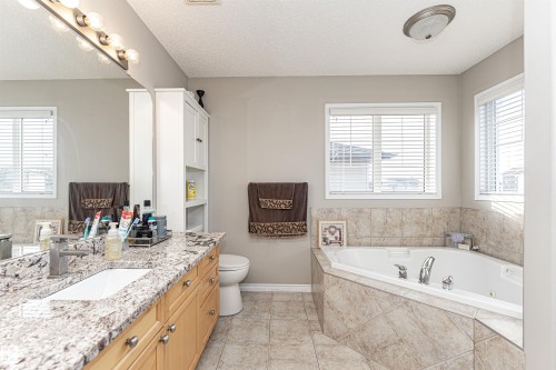 Corner soaking tub with tiled surround, dual windows, long vanity with a solid surface countertop, double sinks, and wood-finish cabinetry - 192 Keyport Circle, Leduc, AB - Indoor Photo Showing Bathroom