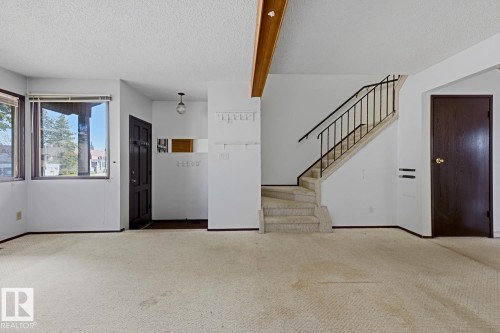 Carpeted flooring, white walls, dark wood-finish entry door, multiple windows with dark frames, and a staircase with black metal railings - 3515 43 Avenue Nw, Edmonton, AB - Indoor Photo Showing Other Room