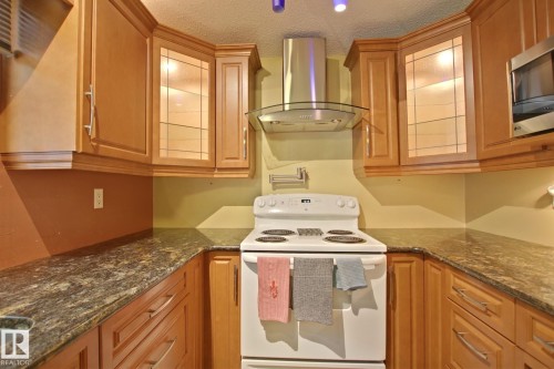 Galley kitchen featuring warm wood cabinetry with glass-front upper cabinets, granite countertops, stainless steel range hood, and white electric range - 18107 89 Avenue Nw, Edmonton, AB - Indoor Photo Showing Kitchen