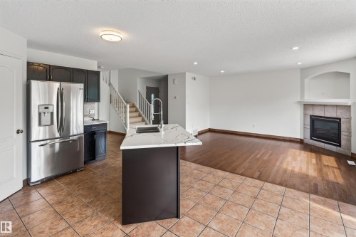 Stainless steel refrigerator, kitchen island with sink and light-toned countertop, dark wood-finish flooring, tiled fireplace with mantel, and recessed lighting - 1707 37A Avenue Nw, Edmonton, AB - Indoor Photo Showing Kitchen