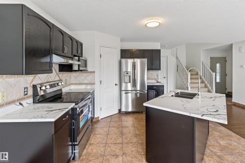 Kitchen featuring dark wood cabinetry, stainless steel appliances, a center island with an undermount sink, white countertops with veining, and a tiled floor - 1707 37A Avenue Nw, Edmonton, AB - Indoor Photo Showing Kitchen With Double Sink