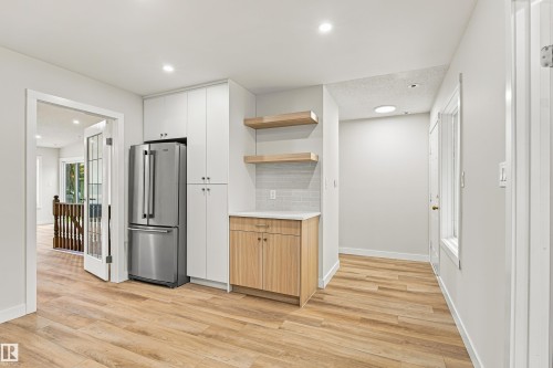 Wood-finish flooring throughout, with white shaker-style cabinetry, a stainless steel refrigerator, light wood-finish lower cabinetry, and open shelving - #21 9375 172 St Nw, Edmonton, AB - Indoor Photo Showing Other Room