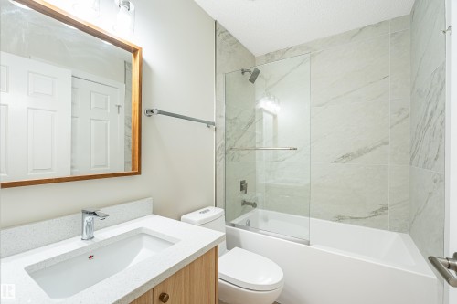 Bathroom featuring a vanity with an integrated sink and light wood-finish cabinetry, a framed mirror, a tub with a glass panel enclosure, and large format wall tile - #21 9375 172 St Nw, Edmonton, AB - Indoor Photo Showing Bathroom