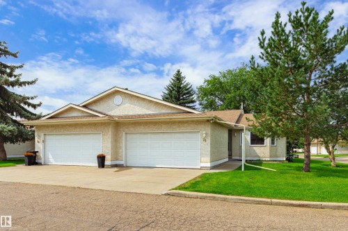 Tan stucco exterior with a brown shingle roof, twin white garage doors, an asphalt driveway, and mature trees - #21 9375 172 St Nw, Edmonton, AB - Outdoor