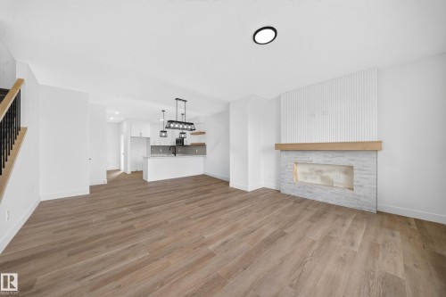 Open-concept living space with wood-finish flooring, a contemporary fireplace with a stone surround, modern kitchen with white cabinetry, and a staircase with a black metal railing - 222 Black Stone Drive Drive, Leduc, AB - Indoor Photo Showing Living Room With Fireplace