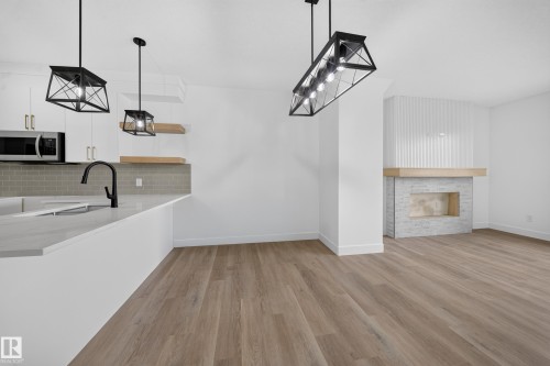 Wood-finish flooring, built-in fireplace with stone surround and wood mantel, white cabinetry with gold-finish hardware, gray subway tile backsplash, and a black-finish gooseneck faucet - 222 Black Stone Drive Drive, Leduc, AB - Indoor With Fireplace