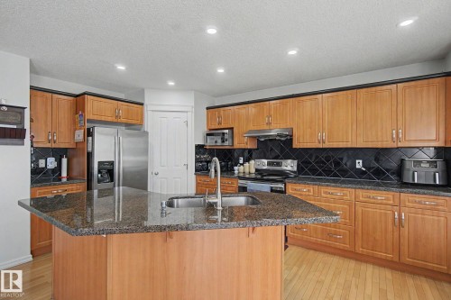 Kitchen featuring wood-finish cabinetry, stainless steel appliances, a dark tile backsplash, and an island with a built-in sink and stone-finish countertop - 3236 21 Street Nw, Edmonton, AB - Indoor Photo Showing Kitchen
