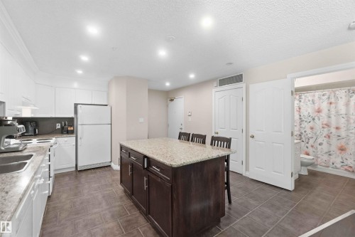 Kitchen featuring white cabinetry, a central island with a stone countertop, a double basin sink, and tile flooring - #104 10221 111 St Nw, Edmonton, AB - Indoor Photo Showing Kitchen