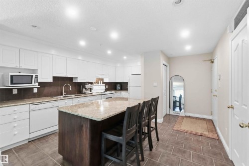 Modern kitchen featuring white cabinetry, a large center island with seating, a dark backsplash, and tile flooring - #104 10221 111 St Nw, Edmonton, AB - Indoor Photo Showing Kitchen With Double Sink