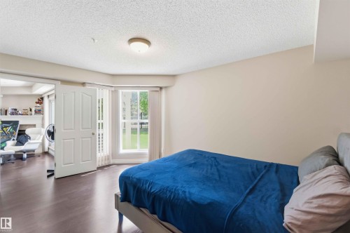 Wood-finish flooring throughout, bay windows with white trim, a large window with exterior landscaping, and a semi-flush mount ceiling light fixture - #104 10221 111 St Nw, Edmonton, AB - Indoor Photo Showing Bedroom