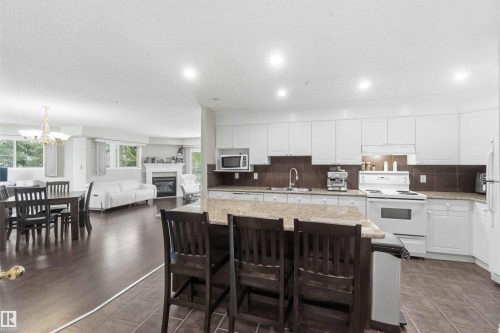 Open-concept kitchen and dining area featuring white cabinetry with quartz countertops, stainless steel appliances, a large center island with seating for three, wood-finish flooring throughout, and recessed lighting - #104 10221 111 St Nw, Edmonton, AB - Indoor